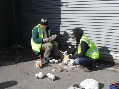 Two technicians in hi-vis vests reviewing paperwork beside rows of soil sample jars