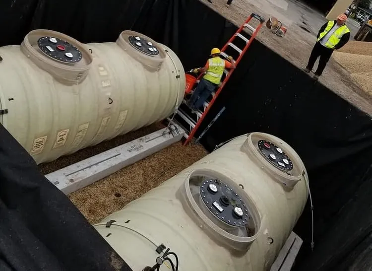 Two new fiberglass underground tanks set in a gravel-lined pit with a worker on a ladder
