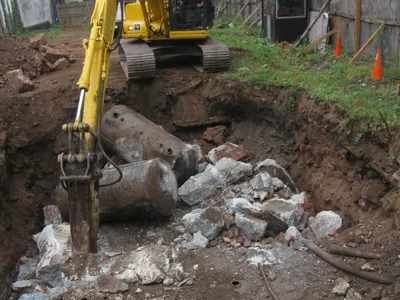 Excavator breaker removing buried steel tanks amid concrete and brick rubble at a brownfield lot