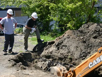 Inspector watching a gloved worker examine excavated soil by a Case backhoe bucket