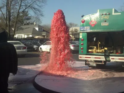 Bright red liquid erupting from the ground beside a green tanker truck in a parking lot