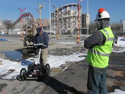 Operator pushing a ground-penetrating radar cart across a lot with a roller coaster behind