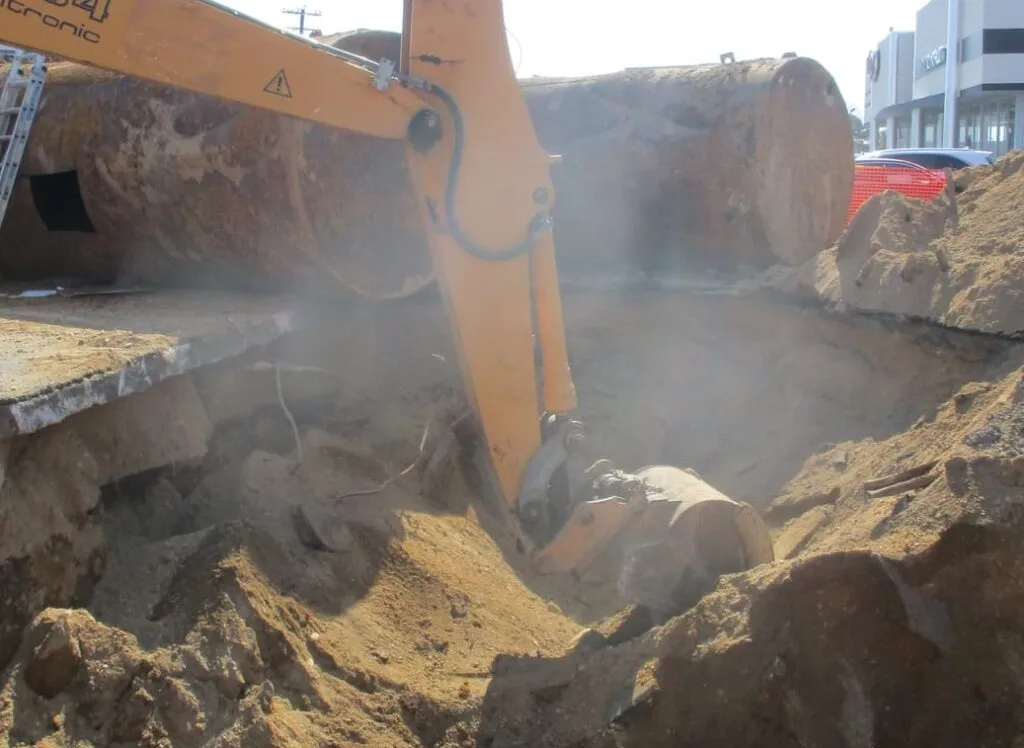 Excavator digging a sandy pit with removed rusty steel tanks lying alongside
