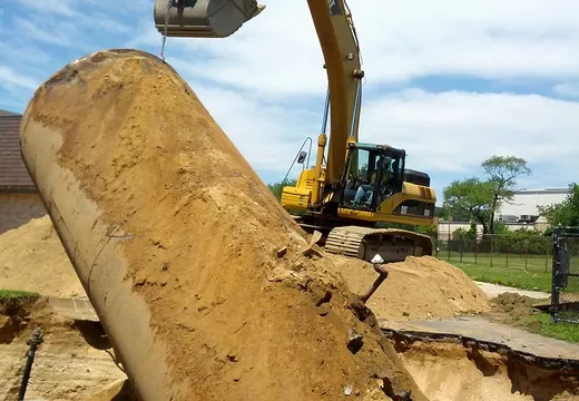 Excavator lifting a steel underground tank out of an excavated pit