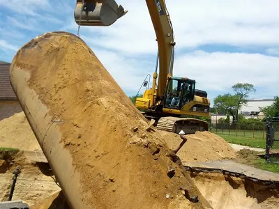 Yellow excavator hoisting a steel tank from an open excavation pit