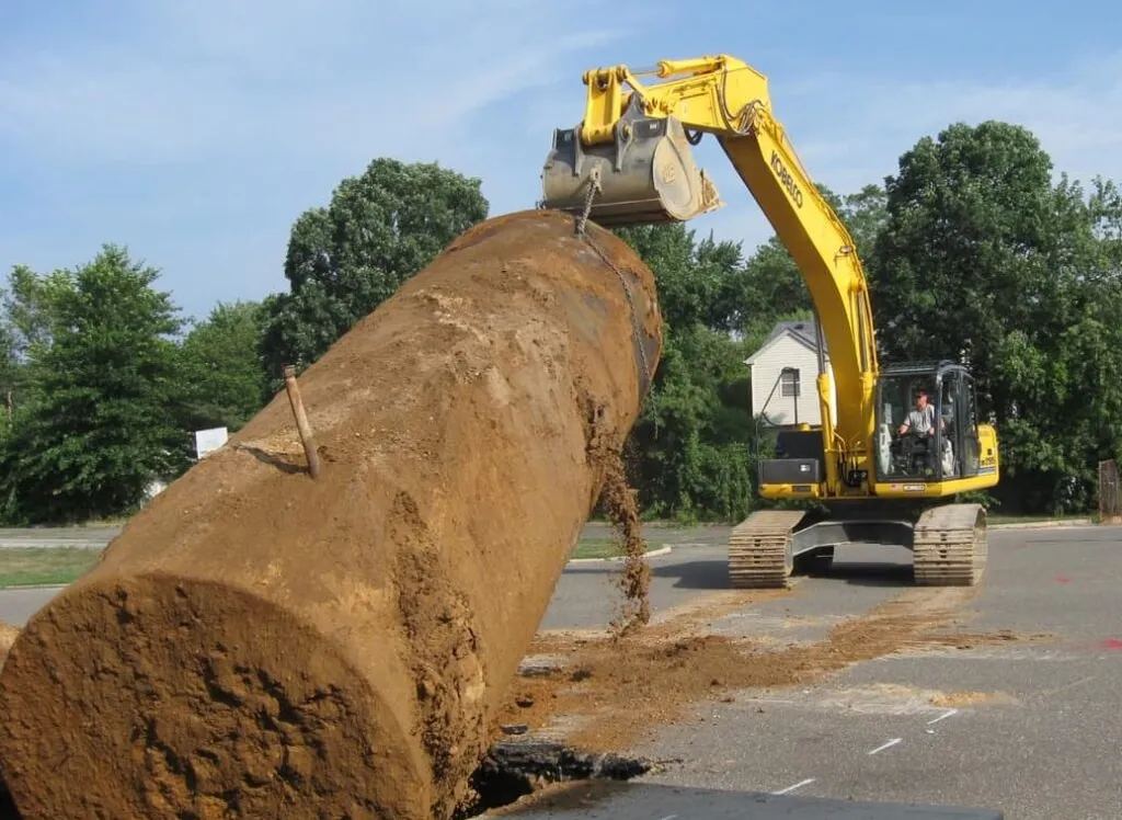 Yellow Kobelco excavator dragging a soil-covered steel underground tank from a parking-lot pit