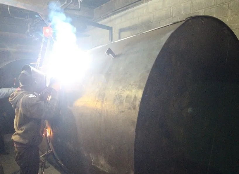Welder fabricating a large cylindrical steel storage tank with a bright welding arc in a basement