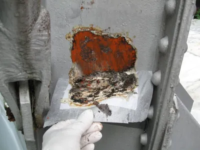 Gloved hand collecting layered paint chips from a riveted steel column