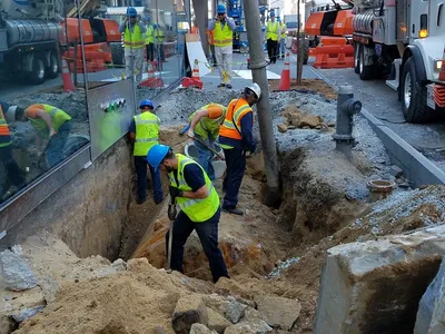 Crew hand-digging an open trench along a city sidewalk excavation