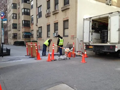 Workers in hi-vis vests coring pavement at a city curb near a box truck with drums