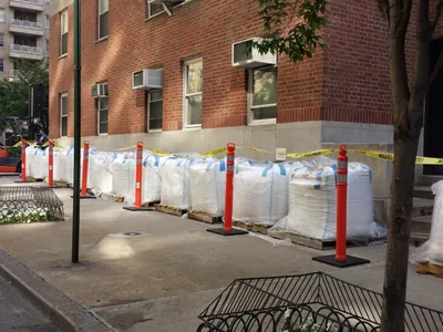 Pallets of shrink-wrapped bulk bags staged behind caution tape on a city sidewalk
