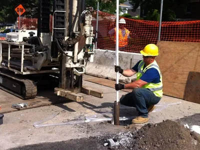Worker in hard hat operating a track-mounted drill rig to install a curbside well