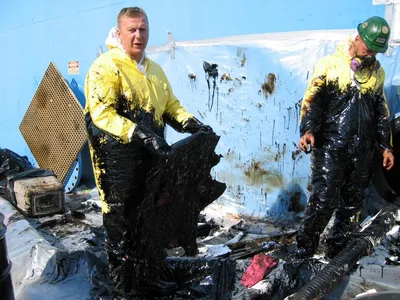 Workers in yellow protective suits removing black sludge from a tank
