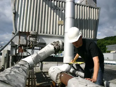 Inspector on a rooftop noting deteriorated insulation on industrial pipes near ductwork