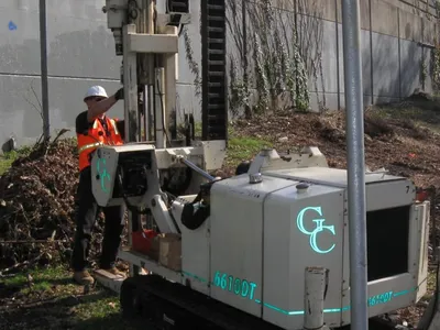 Operator running a tracked Geoprobe 6610DT direct-push rig beside a concrete wall