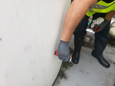 Gloved worker sampling a fitting at the base of a white poly chemical tank