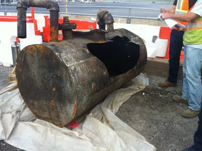 Workers inspecting a corroded steel tank with a rusted-through hole on plastic sheeting