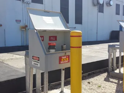 Open solvent tank fill cabinet marked Tank 1 with yellow protective bollards