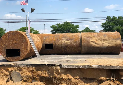 Sectioned rusty steel tank with cut access holes beside an open excavation