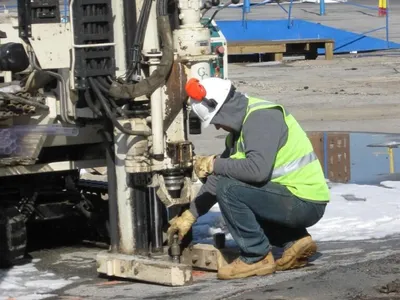 Technician kneeling at the base of a Geoprobe rig on a snowy paved lot
