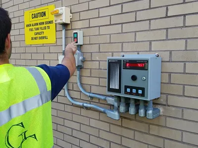 Worker testing a wall-mounted fuel monitor beside an overfill caution sign