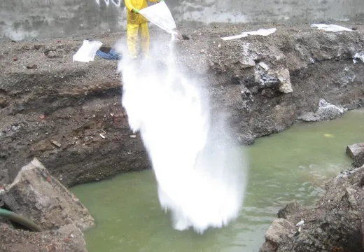 Worker in yellow rain gear pouring white absorbent powder into a water-filled excavation pit