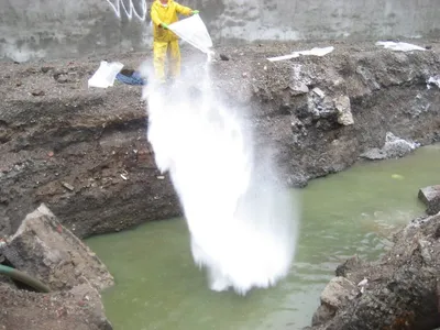 Worker in yellow rain gear pouring white treatment powder into a flooded excavation