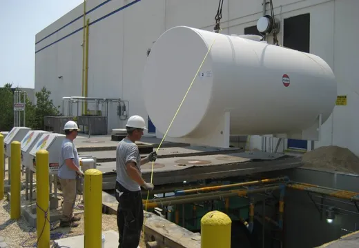 Crane lowering a large white aboveground fuel tank as two workers guide it