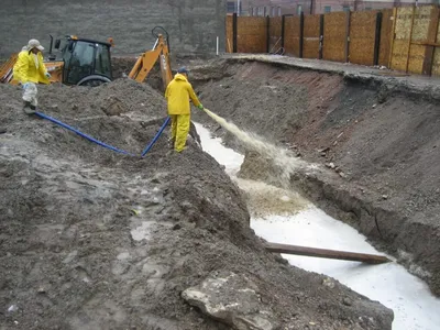 Workers spraying slurry into a lined trench beside a Case backhoe at a fenced site