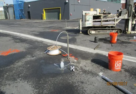 Groundwater wellhead and orange sampling buckets beside a tracked drill rig on pavement