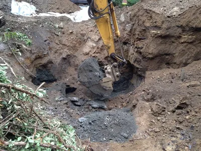 Excavator bucket digging dark contaminated soil from an open excavation pit