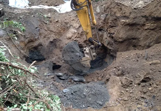Excavator bucket digging dark contaminated soil from an open excavation pit