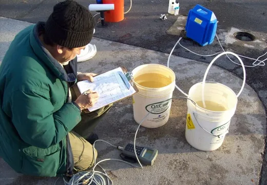 Technician logging groundwater data beside two buckets of turbid water and a field meter