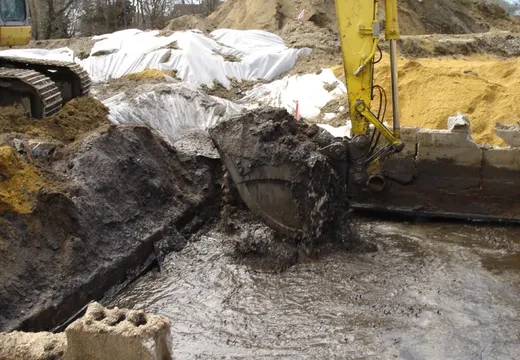 Excavator digging a water-filled trench beside plastic-wrapped soil stockpiles