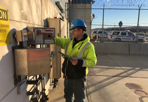 Technician in hard hat checking a high-level tank alarm panel at an airport