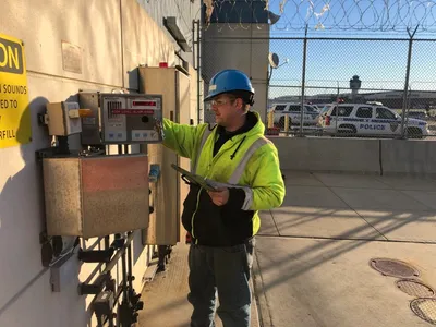 Technician in hard hat checking a high-level tank alarm panel at an airport