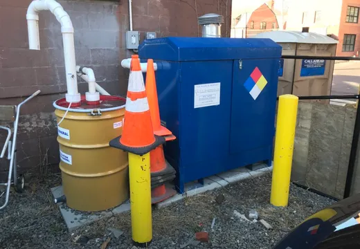 Blue chemical storage cabinet and labeled absorbent drum beside a building exterior