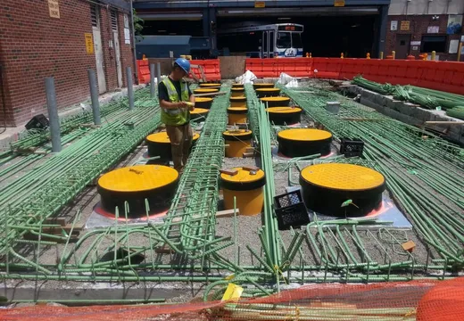 Worker inspecting rows of green piping and yellow sump covers at an urban fuel-system job