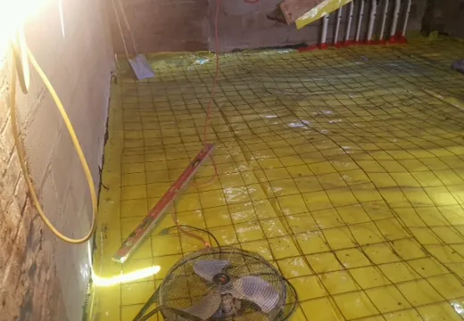 Yellow waterproofing membrane and rebar grid across a basement floor with a drying fan