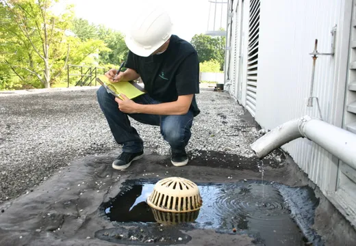 Inspector with a clipboard examining a roof drain surrounded by standing water