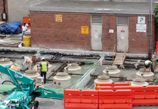 Workers and a crane installing concrete tank access rings behind orange site barriers