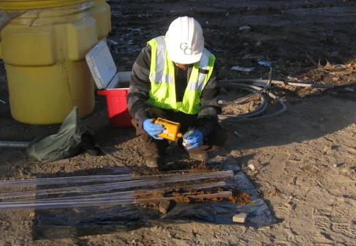 Worker screening soil cores with a handheld meter beside a drum and laid-out samples