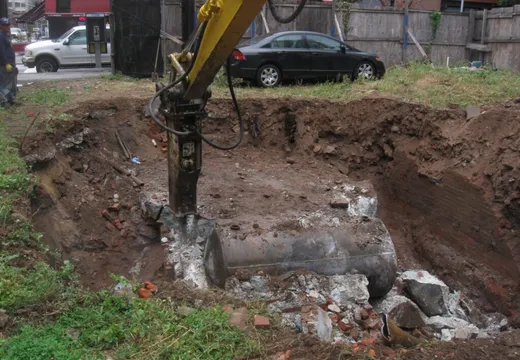 Excavator with hydraulic breaker exposing a buried steel tank in an urban lot
