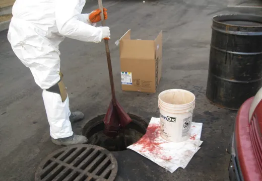 Worker in Tyvek suit hand-pumping spilled liquid from an open street catch basin