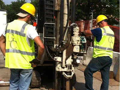 Two G.C. Environmental workers in hi-vis vests operating a direct-push drill rig curbside